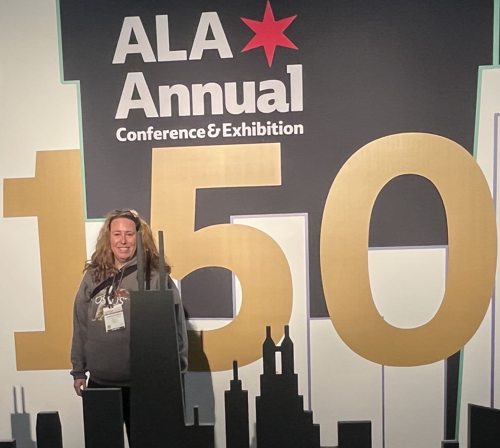 Karin Wannerud, in an OSSUS hoodie and a conference lanyard, standing and smiling at the ALA Annual photo wall in Chicago, in front of the gold 150th-anniversary backdrop with a red star and the city skyline in silhouette.