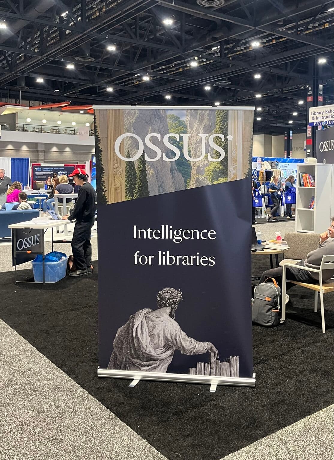 The OSSUS stand on the exhibit floor: a tall roll-up banner reading OSSUS, Intelligence for libraries, with a classical figure illustration, and a staffed table beside it among the aisles.